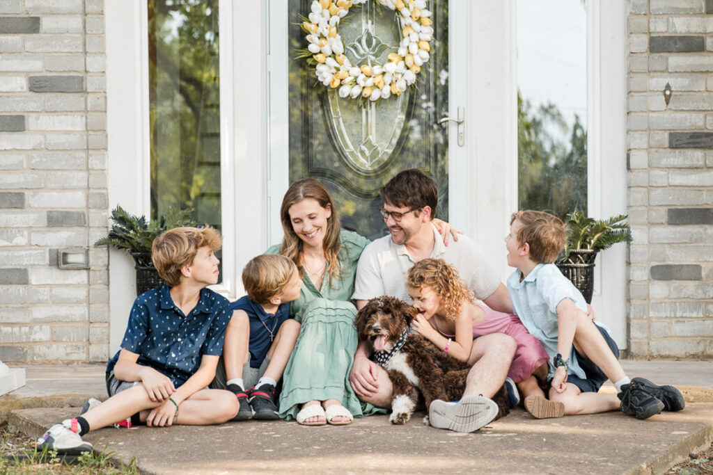 Family of 6 sitting in front of their home snuggling together with their dog.