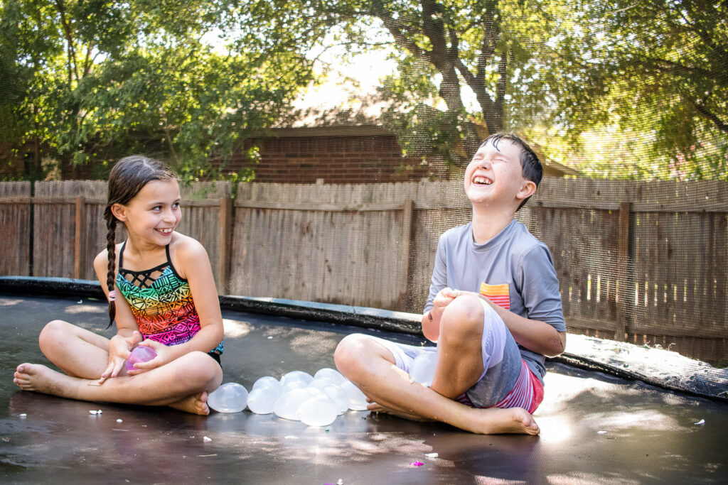 Kids laughing and playing with water balloons on a trampoline during a backyard lifestyle family photography session in Austin, Texas.