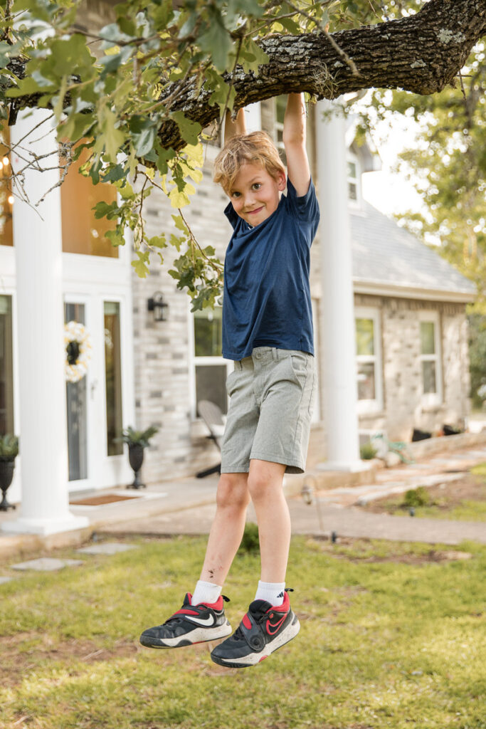Young boy hanging from a tree branch in the yard during a playful lifestyle family photography session in Austin, Texas.