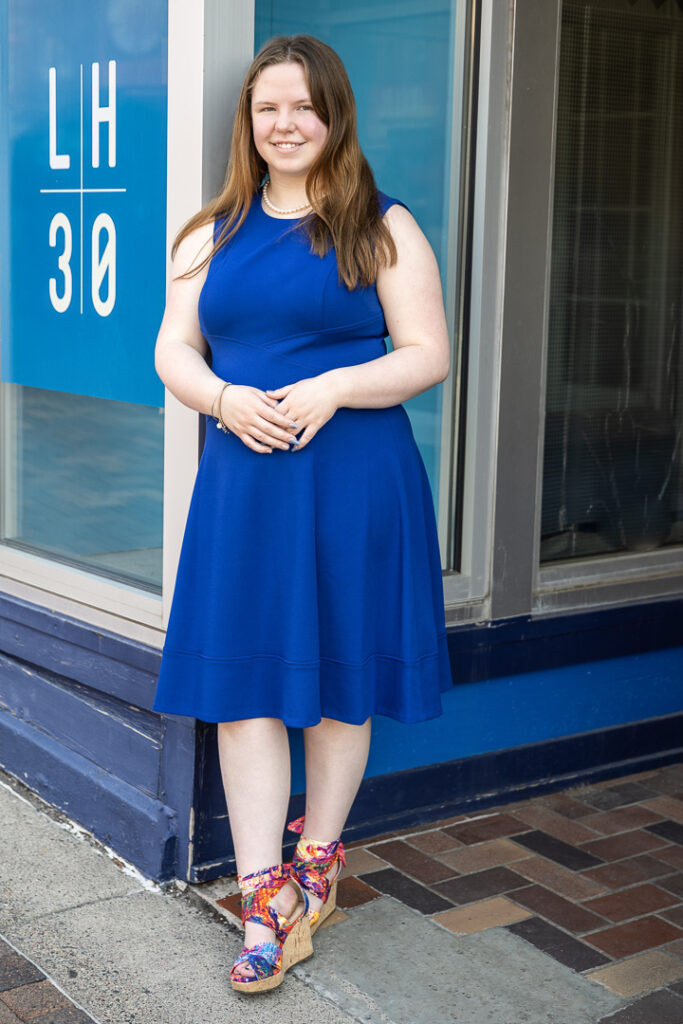 High school senior girl in a blue dress standing outside a colorful storefront in downtown during her senior portraits.