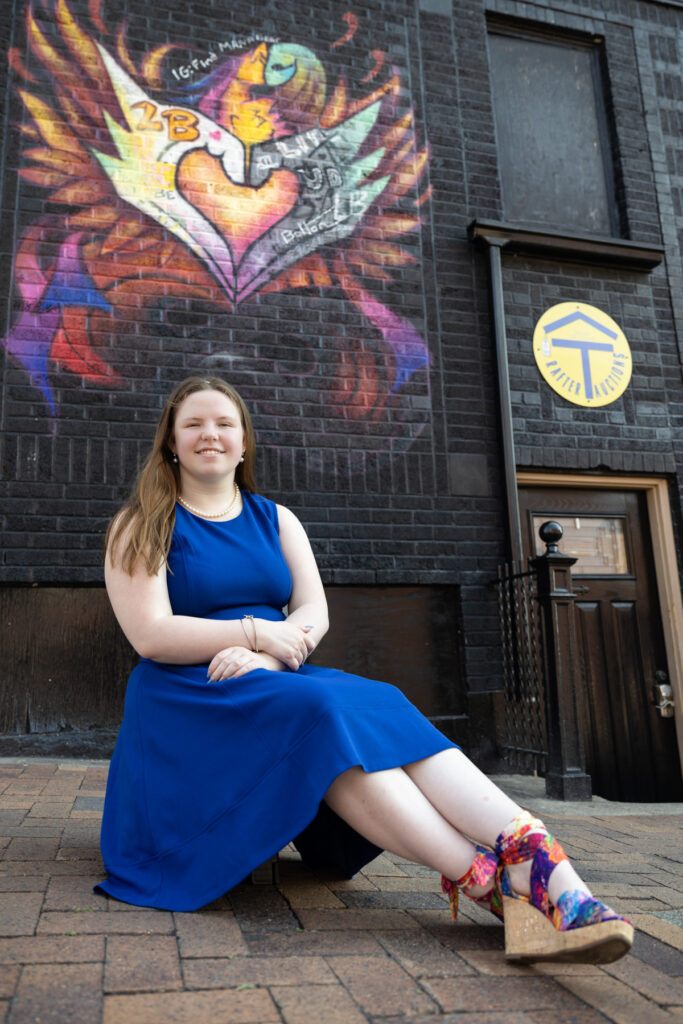 High school senior girl in a blue dress sitting in front of a colorful heart mural.