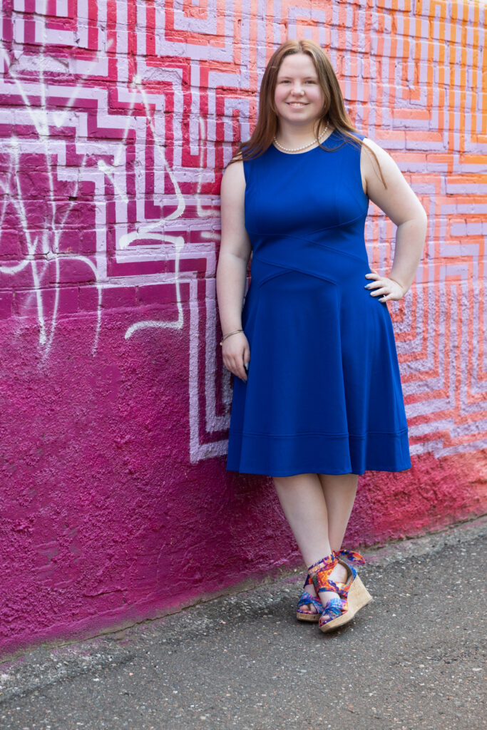 High school senior girl in a blue dress standing outside a colorful storefront during her senior portraits.